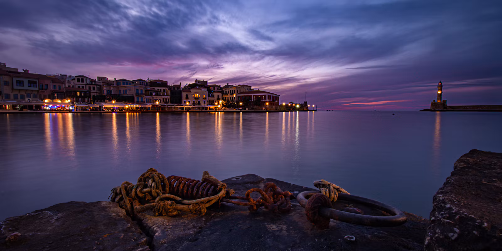 Chania's Venetian harbour at dusk: lit quayside houses on the left, the lighthouse on the right, old mooring chains on the rocks in front