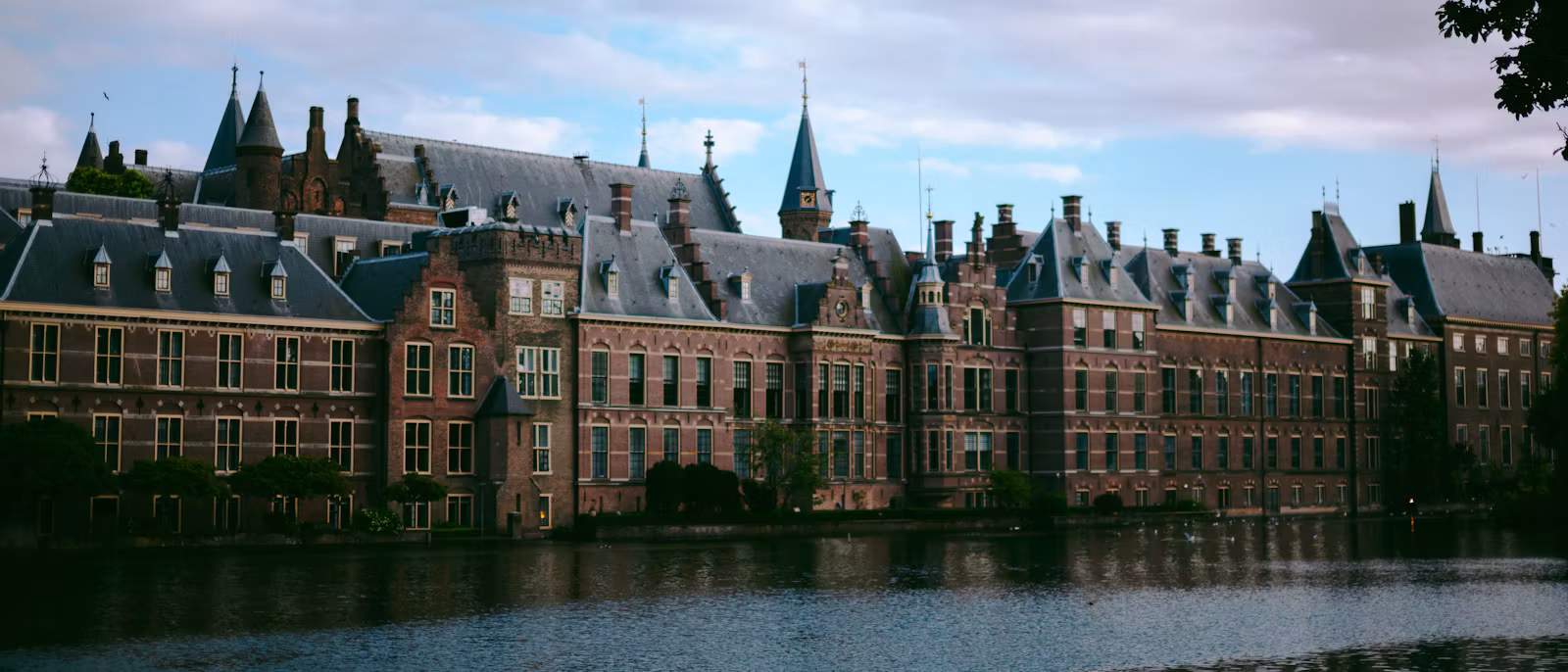 隔着 Hofvijver 湖看国会建筑群 Binnenhof：一长排红砖立面、石板屋顶、塔楼和尖顶倒映在深色的水面上，天上是白云